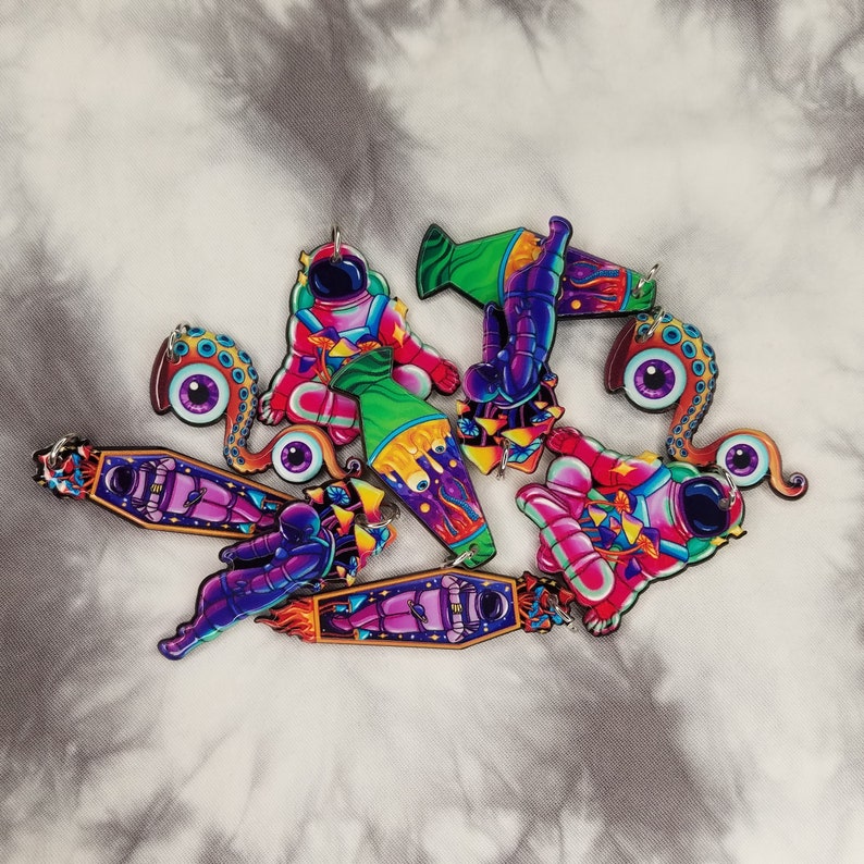 May include: A set of six colorful astronaut and alien themed keychains. The keychains are made of wood and feature intricate details. The keychains are painted in a variety of bright colors, including pink, purple, green, blue, and yellow.