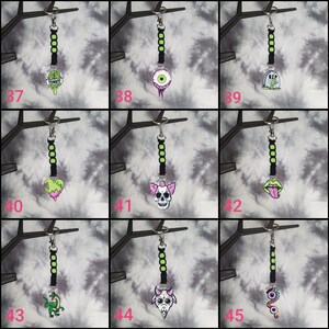 May include: A set of 9 keychains with black and green string and various colorful charms. The charms include a zombie ice cream cone, an eyeball, a tombstone, a bleeding heart, a skull, a tongue, a dinosaur, a goat, and a snake.