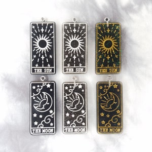 May include: Six black rectangular resin charms with gold and silver glitter. Each charm features a tarot card design with the words "The Sun" and "The Moon" printed on the bottom.