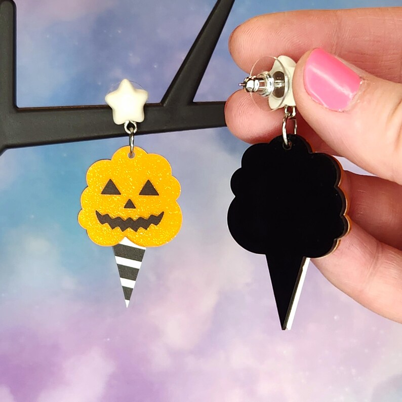 K&ouml;nnte beinhalten: Ein Paar Halloween-Ohrringe mit einem schwarz-wei&szlig; gestreiften Eiswaffelkegel mit einer gelben, k&uuml;rbisf&ouml;rmigen Zuckerwatte oben auf der einen Seite und einer durchgehenden schwarzen Zuckerwatte oben auf der anderen Seite.