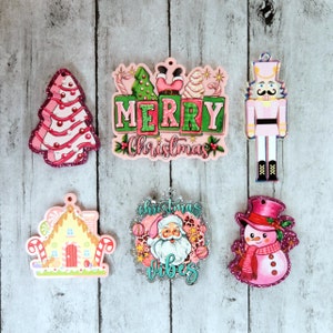Puede incluir: Seis adornos navideños de color rosa y verde con diferentes diseños. Los adornos representan un árbol de Navidad, una casa de jengibre, un muñeco de nieve, un cascanueces, Papá Noel y un cartel que dice "Feliz Navidad".