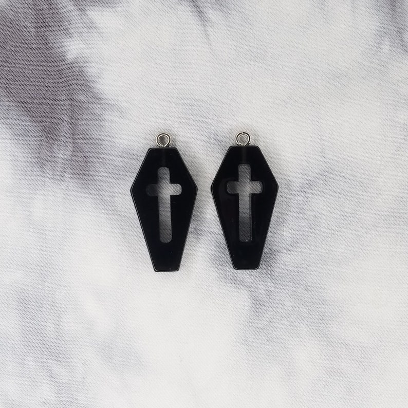 May include: A pair of black coffin-shaped earrings with a cross cut-out. The earrings have silver jump rings.