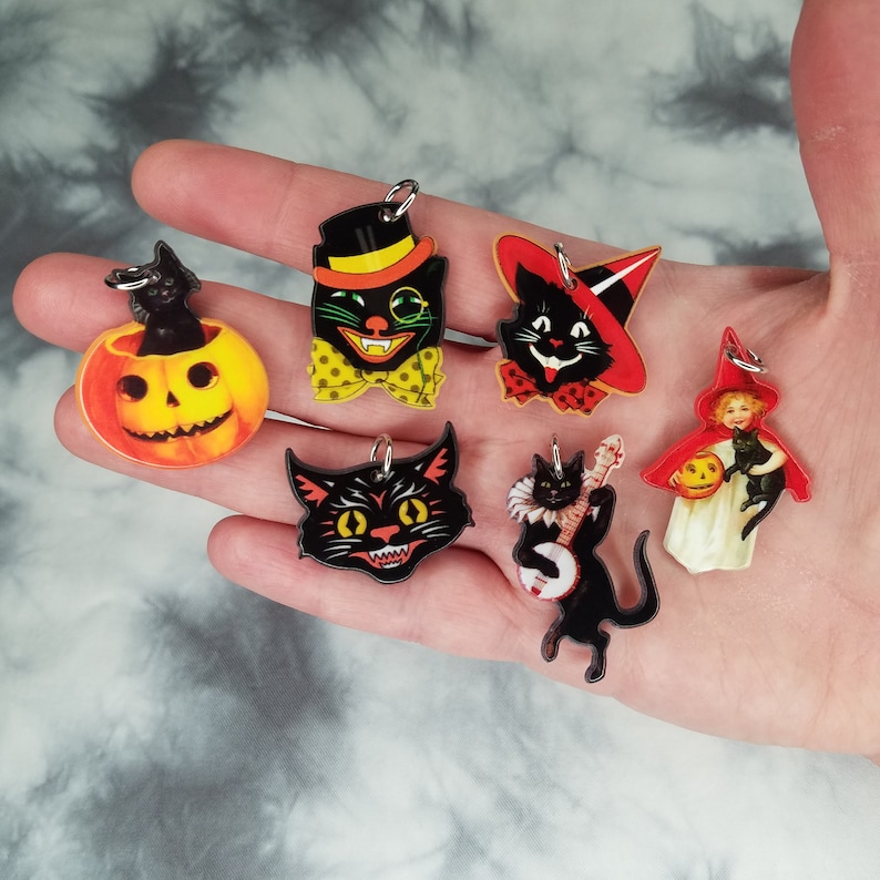 May include: Seven Halloween-themed charms featuring black cats, a jack-o-lantern, and a girl in a red hooded cloak. The charms are made of clear acrylic with vintage-style illustrations.