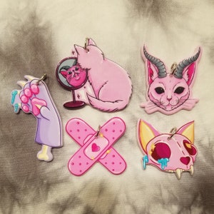 Op de afbeelding: Vijf roze en paarse charmpendants met verschillende ontwerpen, waaronder een kat die in een spiegel kijkt, een kat met hoorns, een pleister, een pootafdruk en een schedel met druipende roze verf.