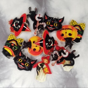 May include: A collection of Halloween-themed ornaments featuring black cats, pumpkins, and witches. The ornaments are made of colorful, painted metal and have a vintage look.