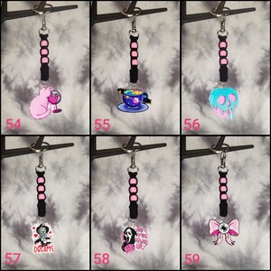 May include: Six different keychains with black and pink cords and various charms. The charms include a pink cat, a blue and purple space-themed teacup, a blue and pink melting skull, a black and white Freddy Krueger character, a white Ghostface character, and a pink bow with an eye.