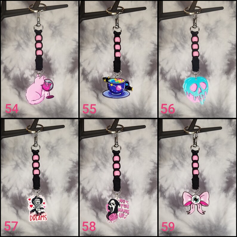 May include: Six different keychains with black and pink cords and various charms. The charms include a pink cat, a blue and purple space-themed teacup, a blue and pink melting skull, a black and white Freddy Krueger character, a white Ghostface character, and a pink bow with an eye.