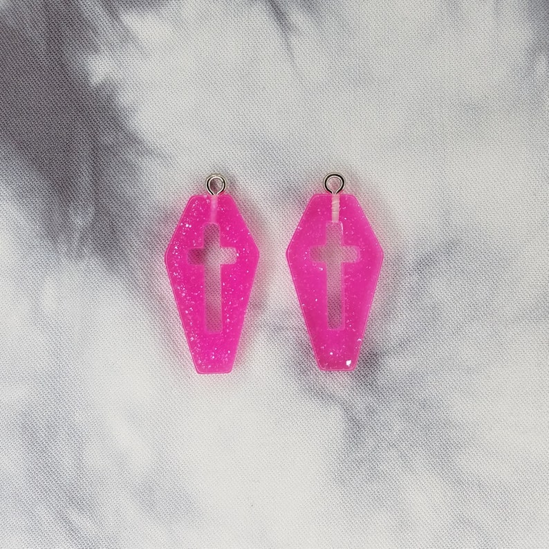 May include: A pair of pink glitter coffin-shaped earrings with a cross cutout.