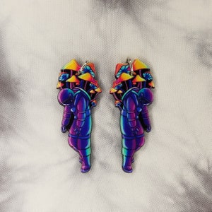 May include: A pair of colorful astronaut earrings with a rainbow gradient and mushroom details. The astronaut is wearing a space suit and has a helmet on. The mushrooms are in a cluster and are also rainbow colored.