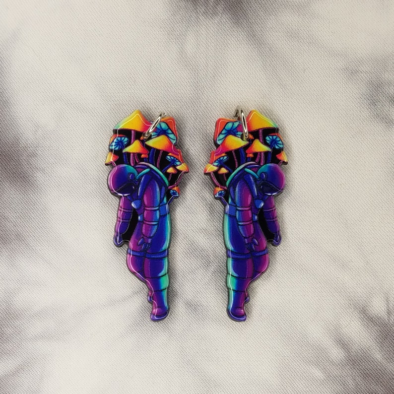 May include: A pair of colorful astronaut earrings with a rainbow gradient and mushroom details. The astronaut is wearing a space suit and has a helmet on. The mushrooms are in a cluster and are also rainbow colored.