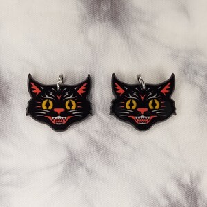 May include: Two black cat face charms with red and yellow details. The charms have a silver loop for hanging.