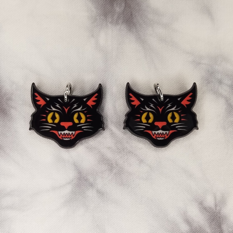 May include: Two black cat face charms with red and yellow details. The charms have a silver loop for hanging.