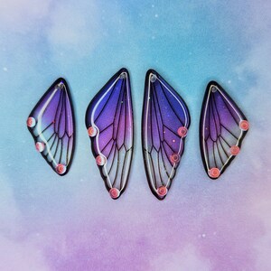Puede incluir: Cuatro pendientes en forma de mariposa de color morado y azul con detalles de purpurina iridiscente. Los pendientes tienen un contorno negro y est&aacute;n acentuados con peque&ntilde;os puntos rosas.