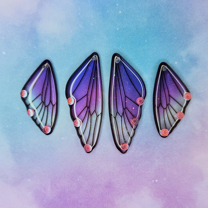 Puede incluir: Cuatro pendientes en forma de mariposa de color morado y azul con detalles de purpurina iridiscente. Los pendientes tienen un contorno negro y est&aacute;n acentuados con peque&ntilde;os puntos rosas.