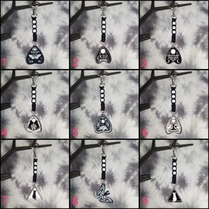 May include: Nine different black and white keychains with a triangular shape and various designs. The designs include a butterfly, a cat, a crescent moon, a pentagram, and the word "Ouija".