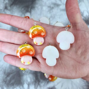 May include: A collection of mushroom-shaped charms. The charms feature orange and yellow caps with white stems. Some have glitter and small dots. Each charm has a silver loop for attaching to jewelry or other crafts.
