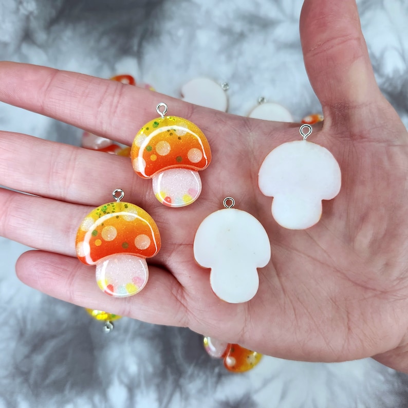 May include: A collection of mushroom-shaped charms. The charms feature orange and yellow caps with white stems. Some have glitter and small dots. Each charm has a silver loop for attaching to jewelry or other crafts.
