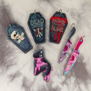 May include: Five different keychains with various designs.  One keychain is a black coffin with the text "Rest in Peace Death" on it. Another keychain is a black coffin with the text "Dead Inside" on it. Another keychain is a red coffin with the text "Home Sweet Home" on it. The last two keychains are knives with pink and red designs.