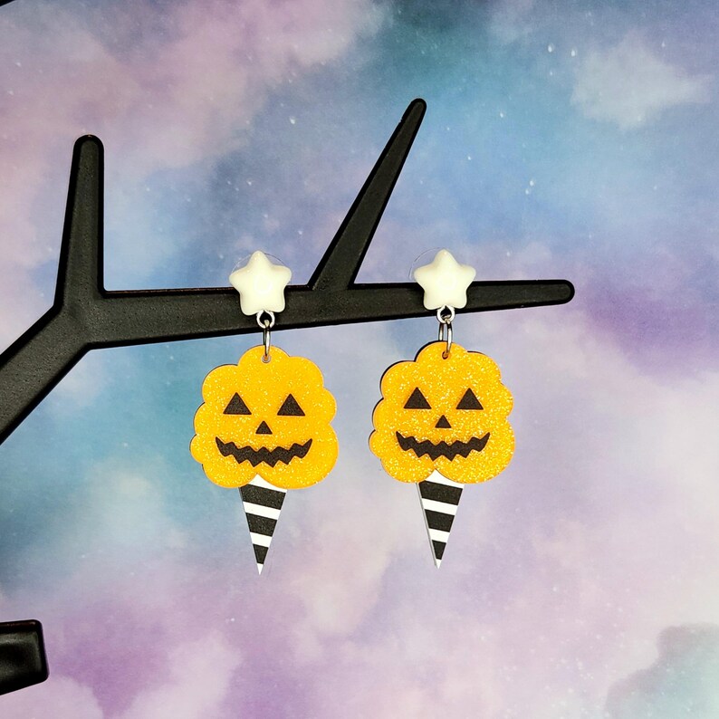 K&ouml;nnte beinhalten: Ein Paar Halloween-Ohrringe mit einem gelben K&uuml;rbis mit einem schwarzen K&uuml;rbisgesicht und einem wei&szlig;en Stern-Anh&auml;nger. Der K&uuml;rbis befindet sich auf einem schwarz-wei&szlig; gestreiften Kegel.