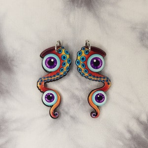 May include: A pair of earrings featuring a colorful octopus design with purple eyes. The octopus is depicted with a swirling, tentacle-like body and a bright orange and yellow color scheme.