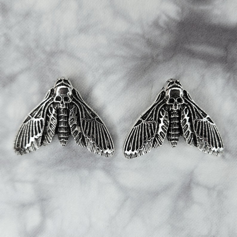 Puede incluir: Un par de pendientes de plata con forma de polilla calavera. Las polillas tienen alas detalladas y un dise&ntilde;o de calavera en el centro.