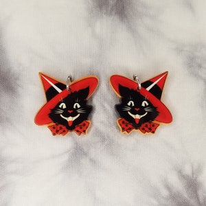 May include: A pair of black cat earrings with red witch hats and white polka dot bows. The earrings are made of acrylic and have a silver metal hook.