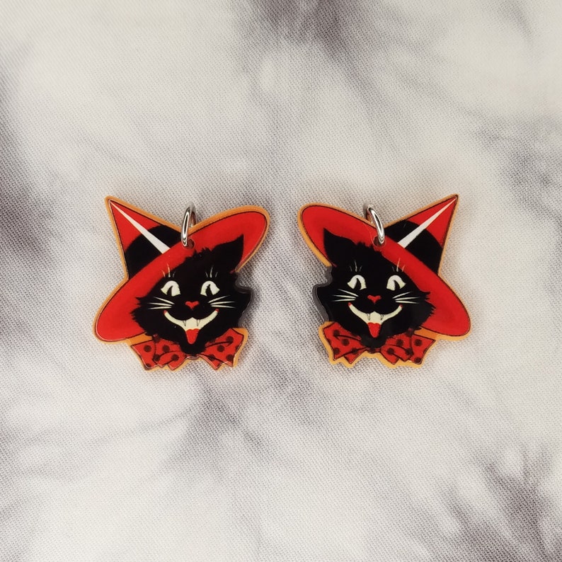 May include: A pair of black cat earrings with red witch hats and white polka dot bows. The earrings are made of acrylic and have a silver metal hook.