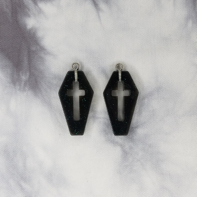 May include: A pair of black coffin-shaped earrings with a silver cross cutout and glitter accents.