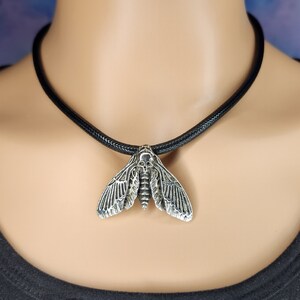 Puede incluir: Un collar con un colgante de polilla de calavera de plata en un cord&oacute;n de cuero negro. La polilla tiene alas detalladas y una cabeza en forma de calavera.