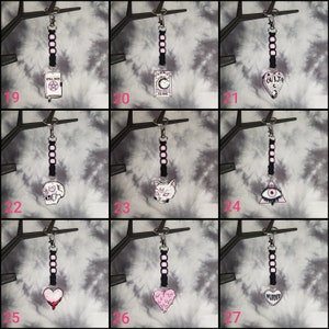May include: Nine black and pink keychains with different charms. The charms include a skull, a cat, a heart, a book, a tarot card, a Ouija board, a triangle with an eye, and a heart with the word "murder".