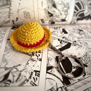 Op de afbeelding: Een gele gehaakte strohoed met een rode band, bevestigd aan een zilveren sleutelhanger. De hoed ligt op een pagina van een manga-stripboek.