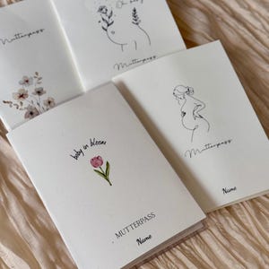 Könnte beinhalten: Vier weiße Mutterpass-Hefte mit Illustrationen und Text. Ein Heft zeigt eine rosa Tulpe und die Worte "baby in bloom". Andere Hefte haben Strichzeichnungen einer schwangeren Person und florale Akzente. Das Wort "Name" ist ebenfalls sichtbar.