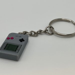 May include: A grey keychain shaped like a retro handheld video game console with pink buttons and a black directional pad.