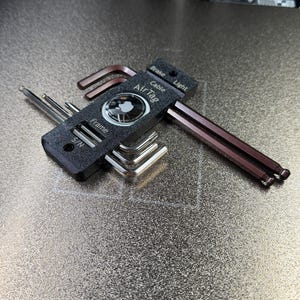 May include: A set of Allen wrenches and an AirTag device. The wrenches are silver and brown, with a black holder. The AirTag is black and white, with the text "AirTag" visible. The tools are on a textured, metallic surface.