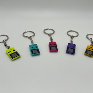 以下が含まれることがあります： 異なる色のゲームコンソールチャームが付いたキーホルダーが5つあります。チャームは、画面、ボタン、方向パッドが付いた携帯用ゲームコンソールのような形をしています。キーホルダーは銀色で、上部に円形のリングが付いています。チャームは黄色、青、ピンク、紫、オレンジです。