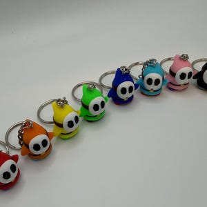 Peut inclure: Une collection de porte-clés Shy Guy colorés. Chaque porte-clés représente un personnage Shy Guy de couleur différente, notamment rouge, orange, jaune, vert, bleu, turquoise, rose et noir. Les porte-clés ont des anneaux argentés.