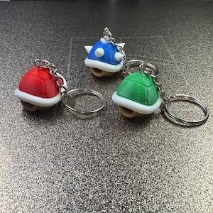 May include: Three turtle shell keychains in red, blue, and green. Each shell features a white rim and a silver metal keyring attached to the top. The blue shell has white spikes on its surface, while the red and green shells have smooth tops.