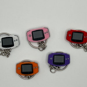 May include: Five different colored keychain charms shaped like a handheld video game console. The charms are white, pink, red, purple, and orange.