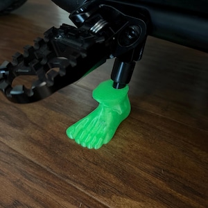 May include: A bright green, 3D-printed foot-shaped kickstand puck attached to the bottom of a black metal motorcycle kickstand. The textured, translucent green plastic foot provides a wider base for the kickstand on the floor surface.