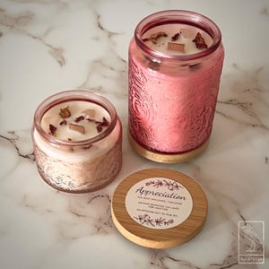 May include: Two pink glass jar candles with decorative etched patterns. The candles have a light pink wax and are topped with dried rose petals and wood pieces. A wooden lid with the word "Appreciation" is next to the candles.