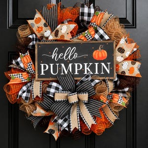May include: A decorative wreath featuring orange, black, and tan mesh ribbons, black and white buffalo check fabric, and tan ribbons with pumpkin prints. A central rectangular sign displays the text hello PUMPKIN with a small orange pumpkin graphic.