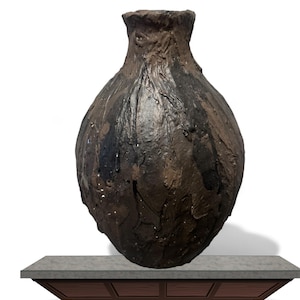 May include: A textured, dark brown ceramic vase with a bulbous body and a rough, uneven surface. The vase has dark, almost black, patches. It sits on a grey countertop with a brown wooden base.