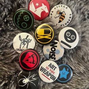 Może przedstawiać: Zbiór 10 okrągłych znaczków z różnymi wzorami. Znaczek są w różnych kolorach, w tym czarny, biały, czerwony, zielony, niebieski, żółty i brązowy. Niektóre z wzorów obejmują kota, pająka, czaszkę, pszczołę, gwiazdę i uśmiechniętą buźkę. Znaczek mają średnicę około 2,5 cm.