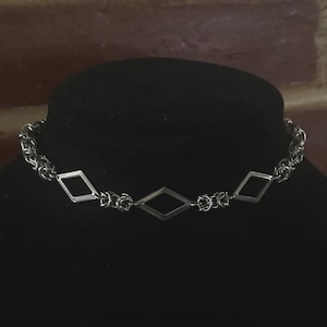 May include: A silver-colored choker necklace with a chainmail design. The necklace features three diamond-shaped pendants. The necklace is displayed on a black mannequin head against a brick background.