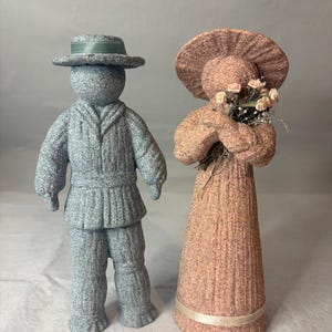 May include: Two textured figurines. The left figure is a blue person wearing a hat with a green ribbon, a collared jacket, and trousers. The right figure is a peach-colored person wearing a wide-brimmed hat and a long dress, holding a bouquet of small flowers.
