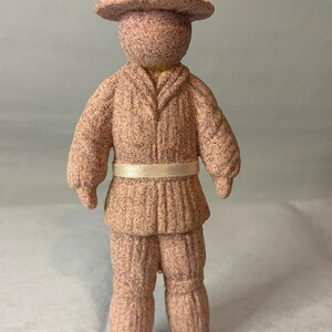 May include: A textured, light brown, speckled figurine of a person wearing a wide-brimmed hat and a matching jacket with a light beige ribbon belt. The figure has a simple, rounded head and long pants, all crafted from a uniform, grainy material.