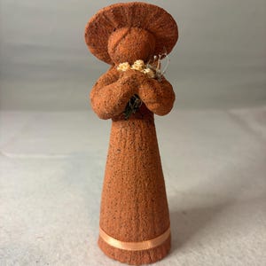 May include: A terracotta-colored figurine with a textured, granular surface. The figure wears a wide-brimmed hat and holds a small cluster of light-colored dried flowers near the chest. A thin, smooth peach-colored ribbon encircles the base.