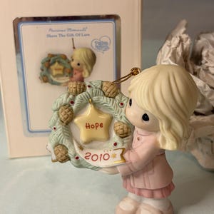 May include: A Precious Moments porcelain figurine of a child with blonde hair wearing a pink coat, holding a green wreath decorated with pinecones and red berries. A yellow star in the center reads Hope, and a white banner below reads 2010.