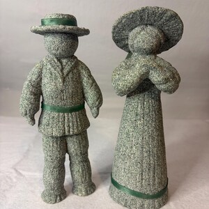May include: Two textured gray stone-like figurines standing side by side. The left figure wears a hat and jacket with a green ribbon. The right figure wears a wide-brimmed hat and a long dress with a green ribbon at the hem. Both have a speckled finish.