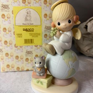 May include: A porcelain figurine featuring a blonde angel with a halo sitting atop a globe, holding a cluster of green grapes. A small gray cat with a heart collar sits on two stacked boxes beside the globe, all on a round base.